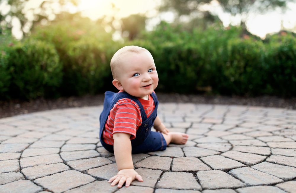 Happy baby in overalls sits on circular stone patio in garden.