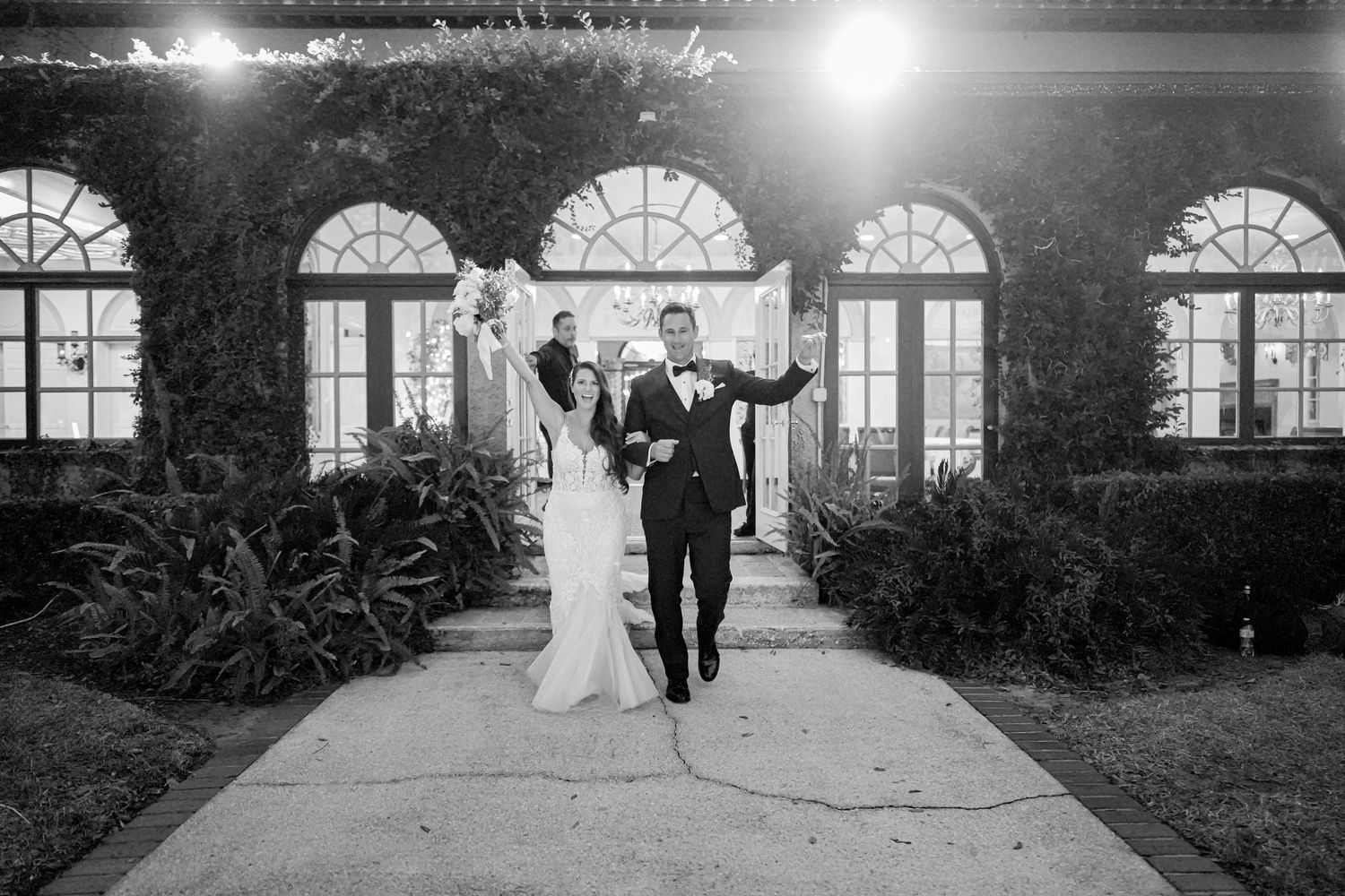 A joyful couple exits an elegant venue through arched French doors illuminated by evening lights and bordered by ferns.
