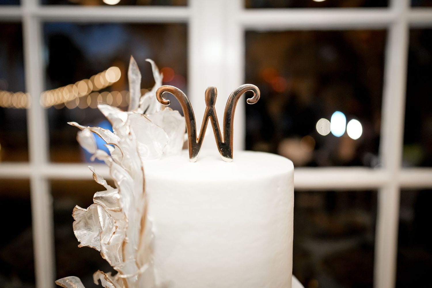 An elegant white wedding cake topped with a gold monogram W against a window backdrop.