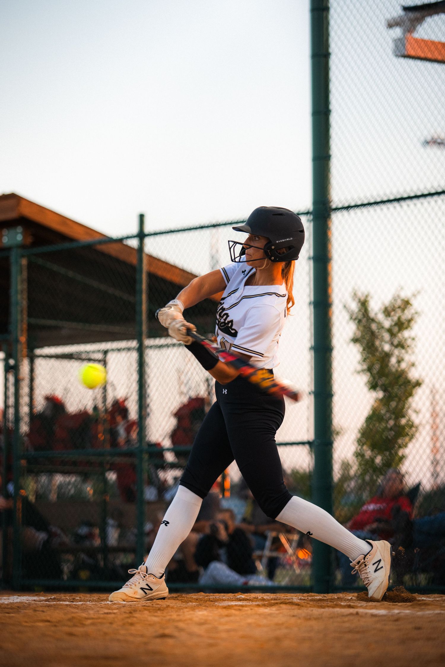 Softball player in white uniform swinging bat at incoming yellow ball on dirt field.