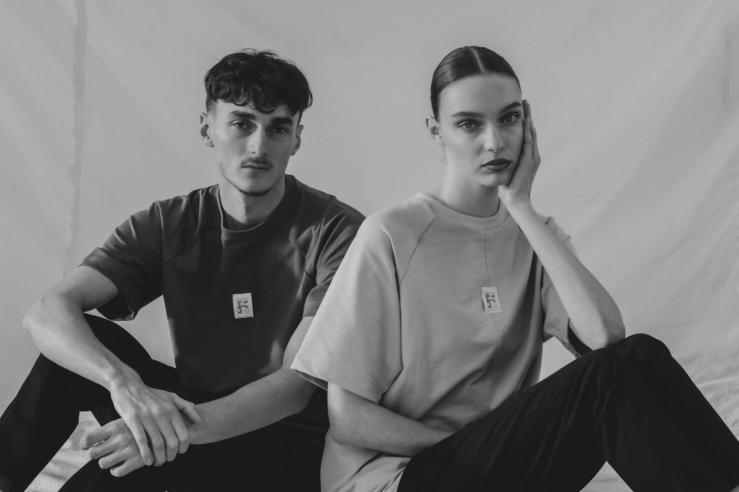 Zwart-wit portret van twee jonge modellen in casual kleding, zittend in een studio setting met minimalistische achtergrond.