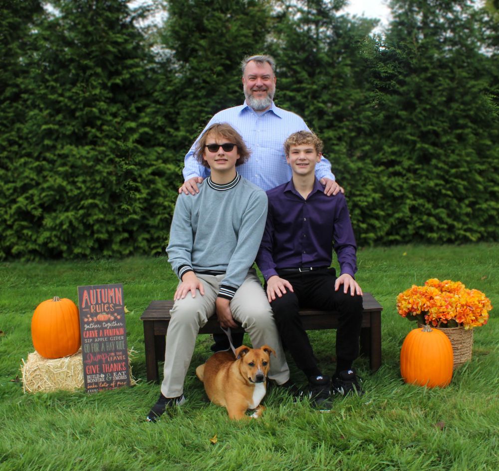 Three people and a Corgi dog pose on bench with autumn decorations and pumpkins.