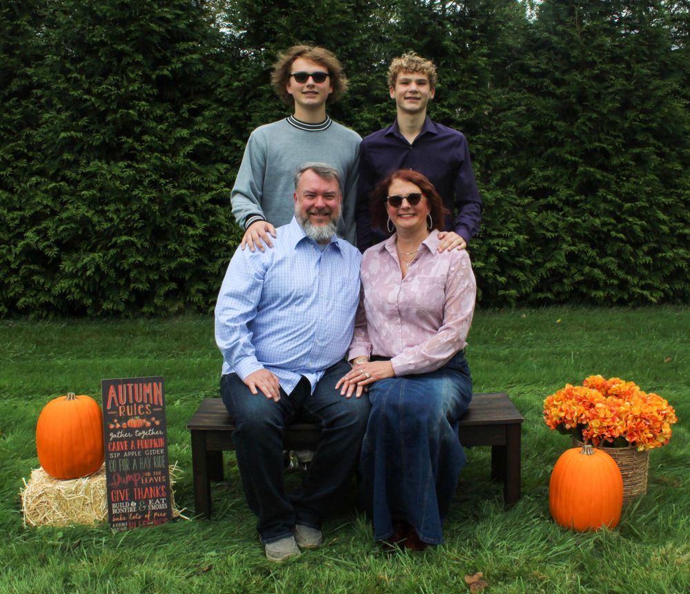 Family photo on garden bench with autumn decorations against green hedge background.