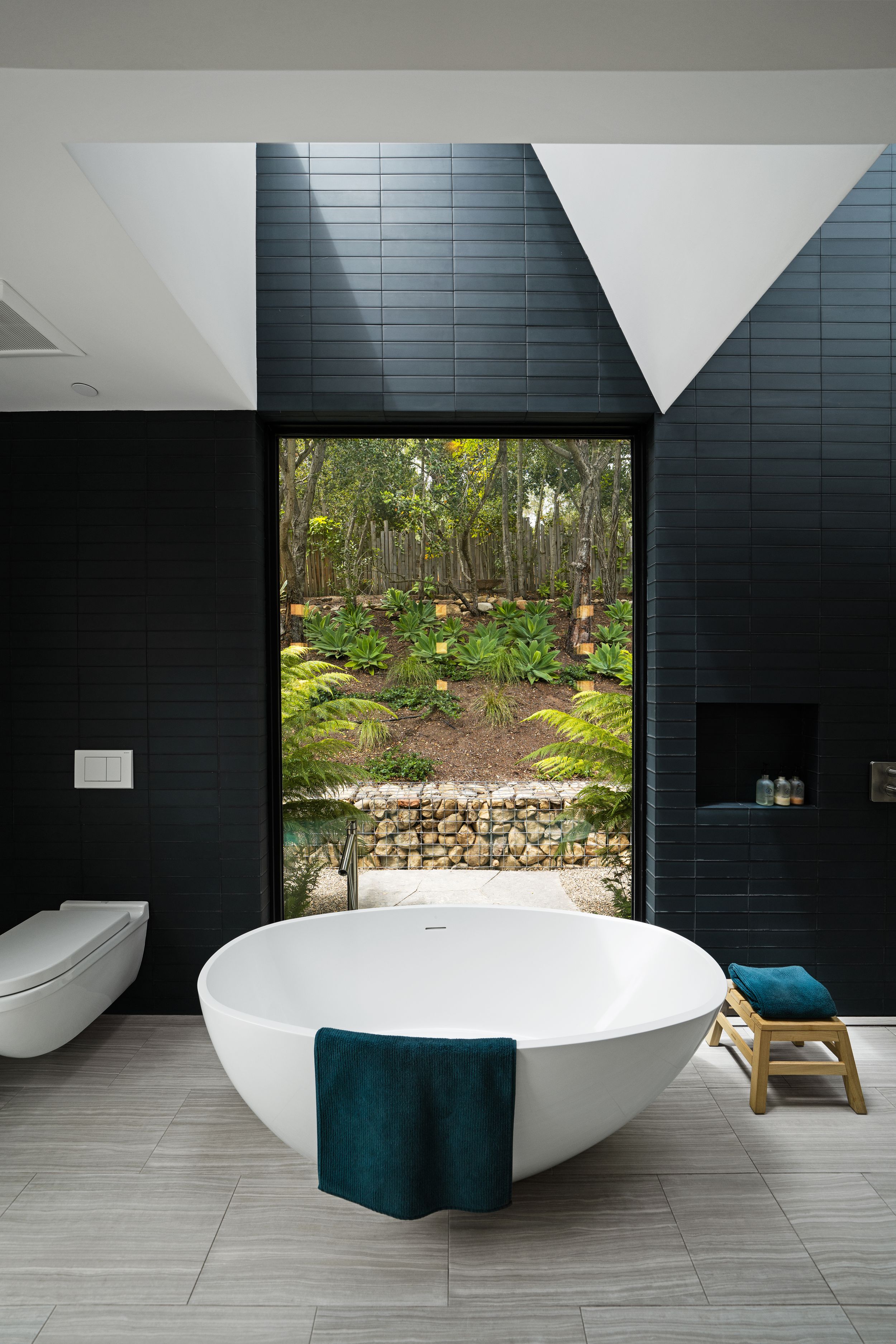 Luxurious bathroom featuring freestanding white tub and black tile walls with large window overlooking garden.