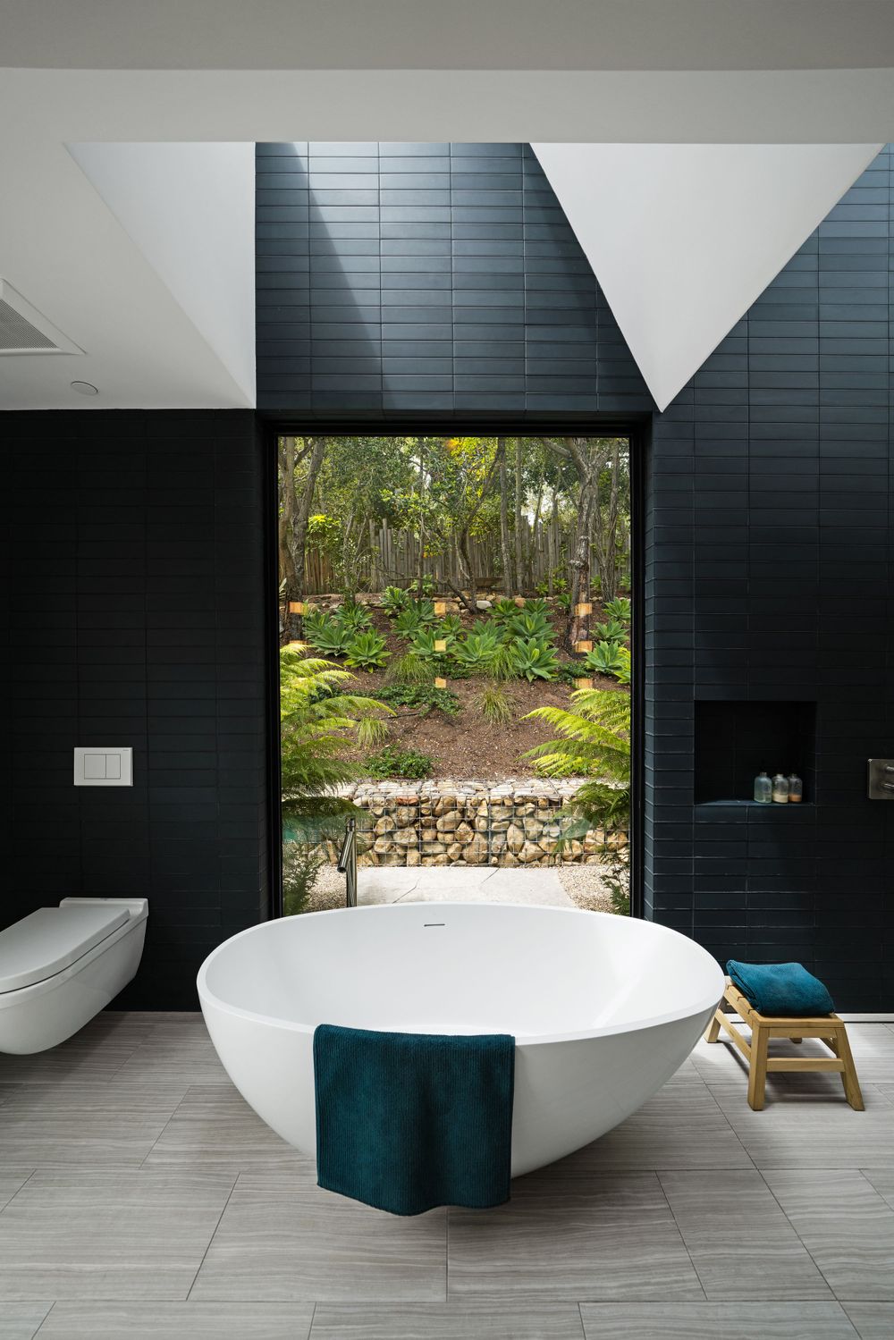 Luxurious bathroom featuring freestanding white tub and black tile walls with large window overlooking garden.