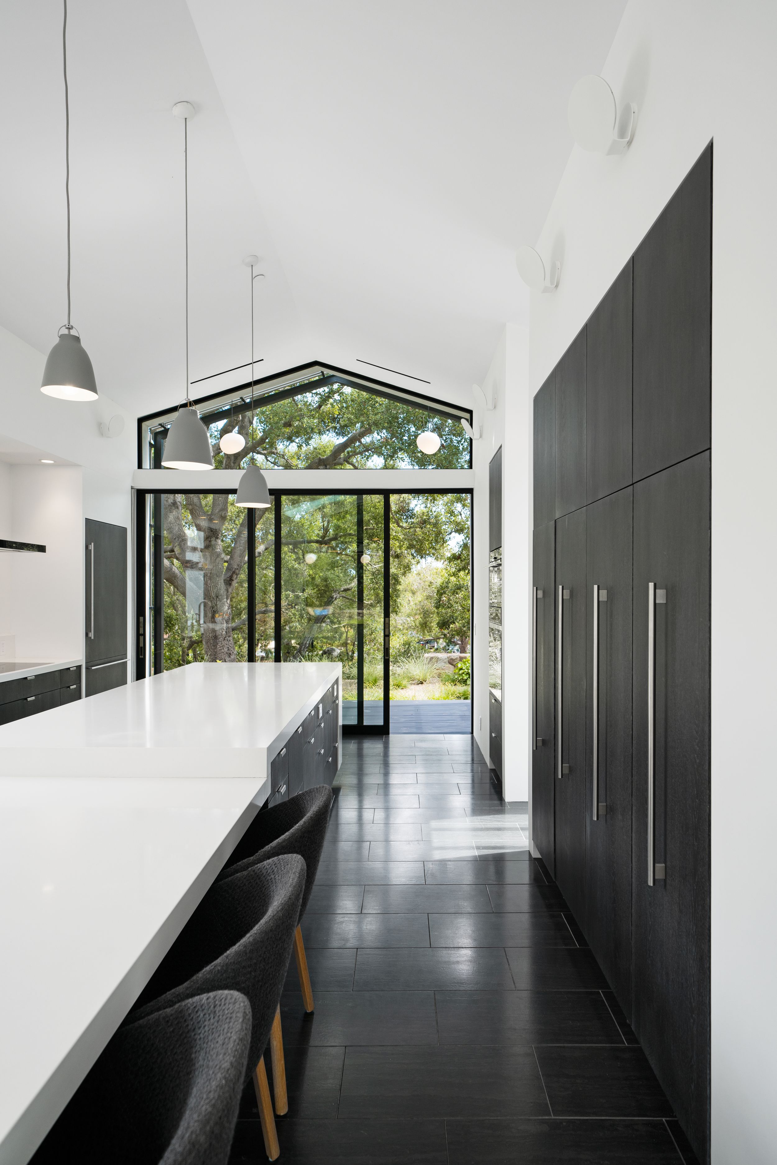 Modern kitchen with black cabinetry, white countertops and dramatic floor-to-ceiling windows framing forest views.