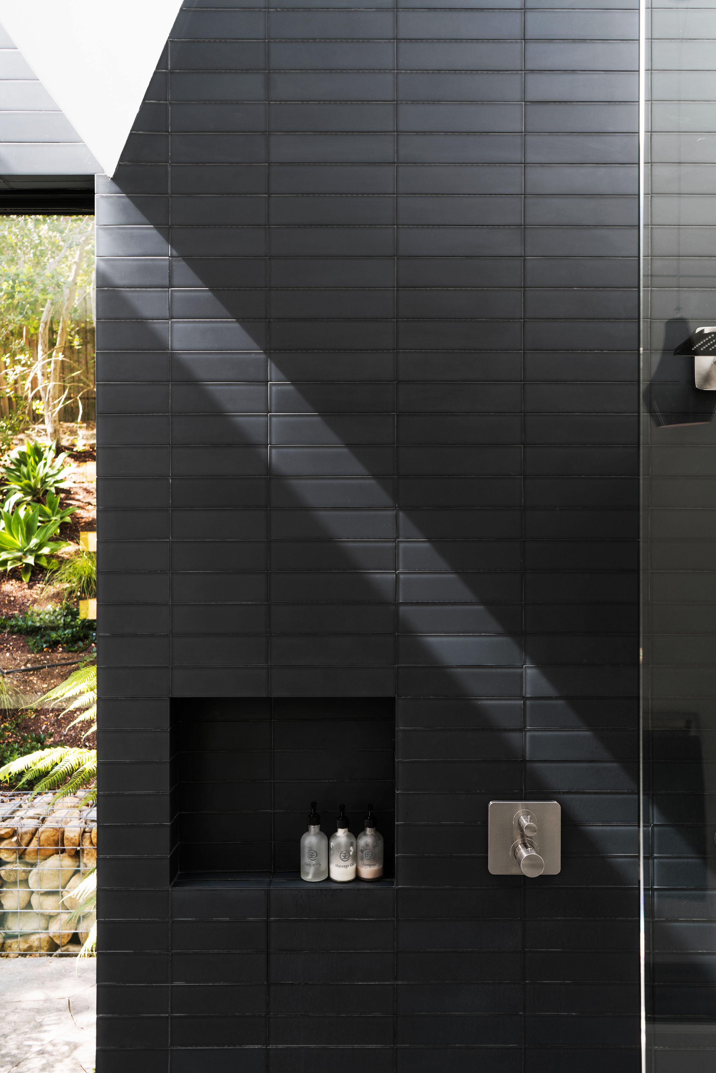 Black tiled exterior wall with built-in shower niche and modern fixtures catching dramatic diagonal sunlight.