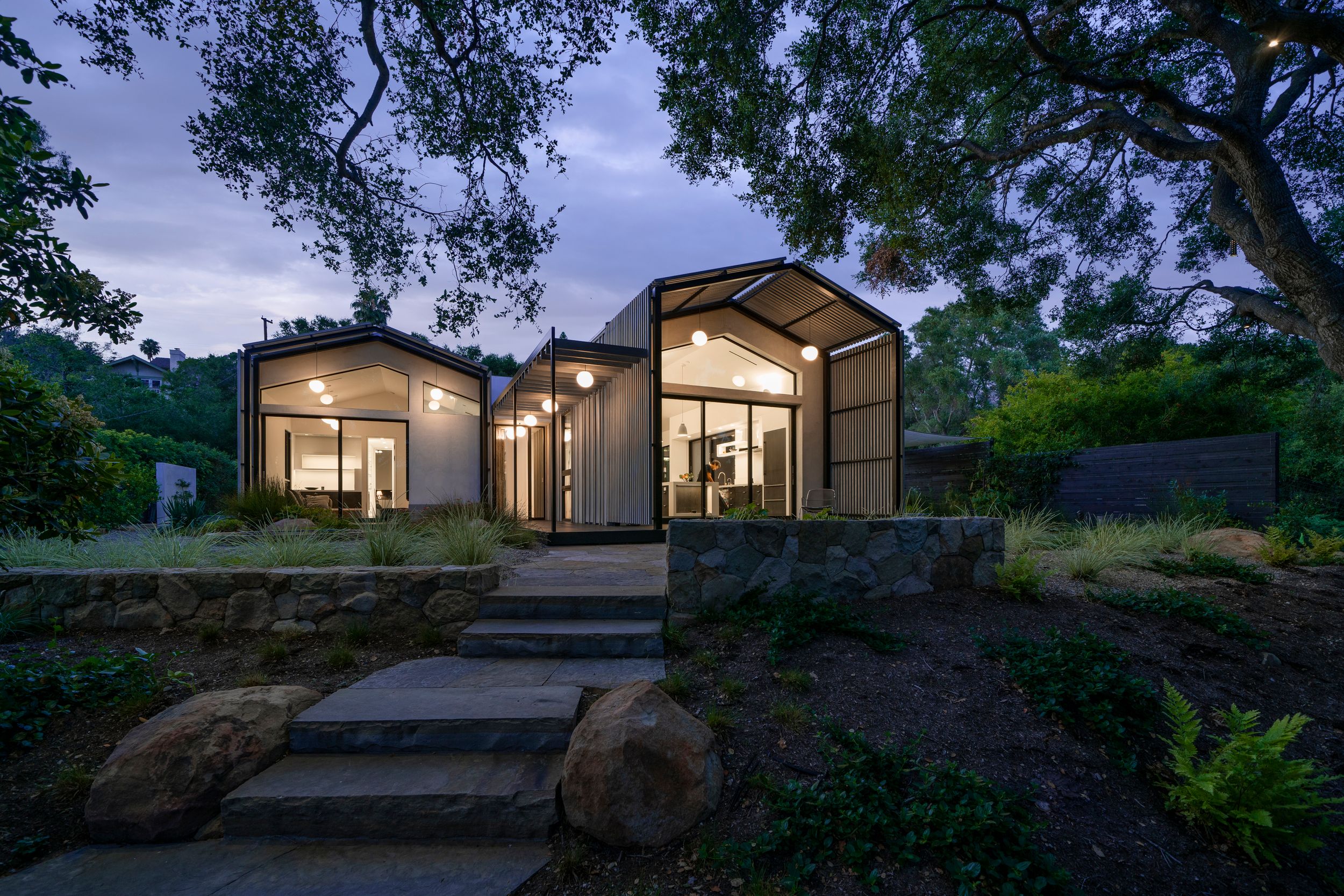 Modern hillside home with glass walls illuminated at dusk among trees.