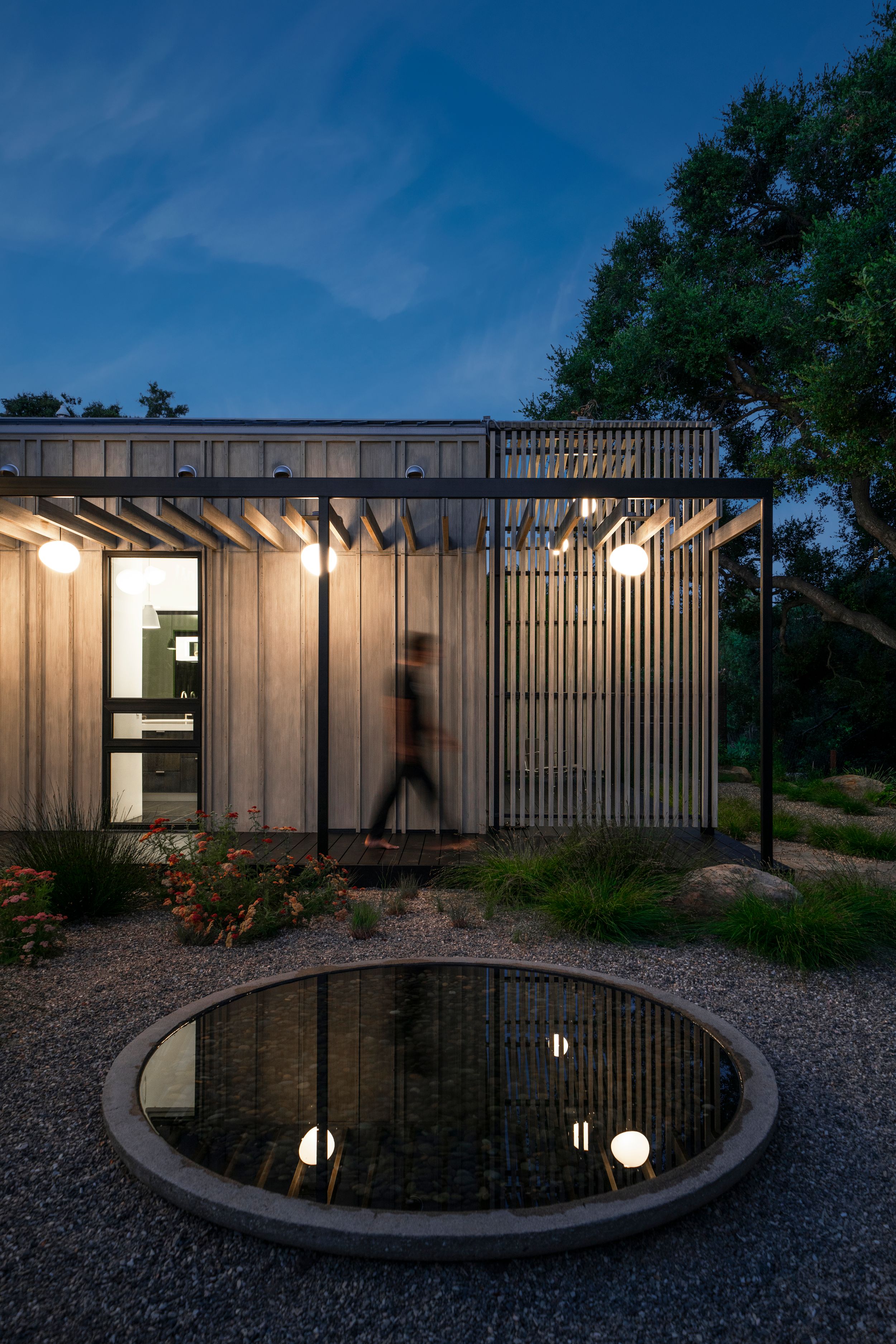 Modern architectural design featuring vertical wood slats and circular reflecting pool at night.