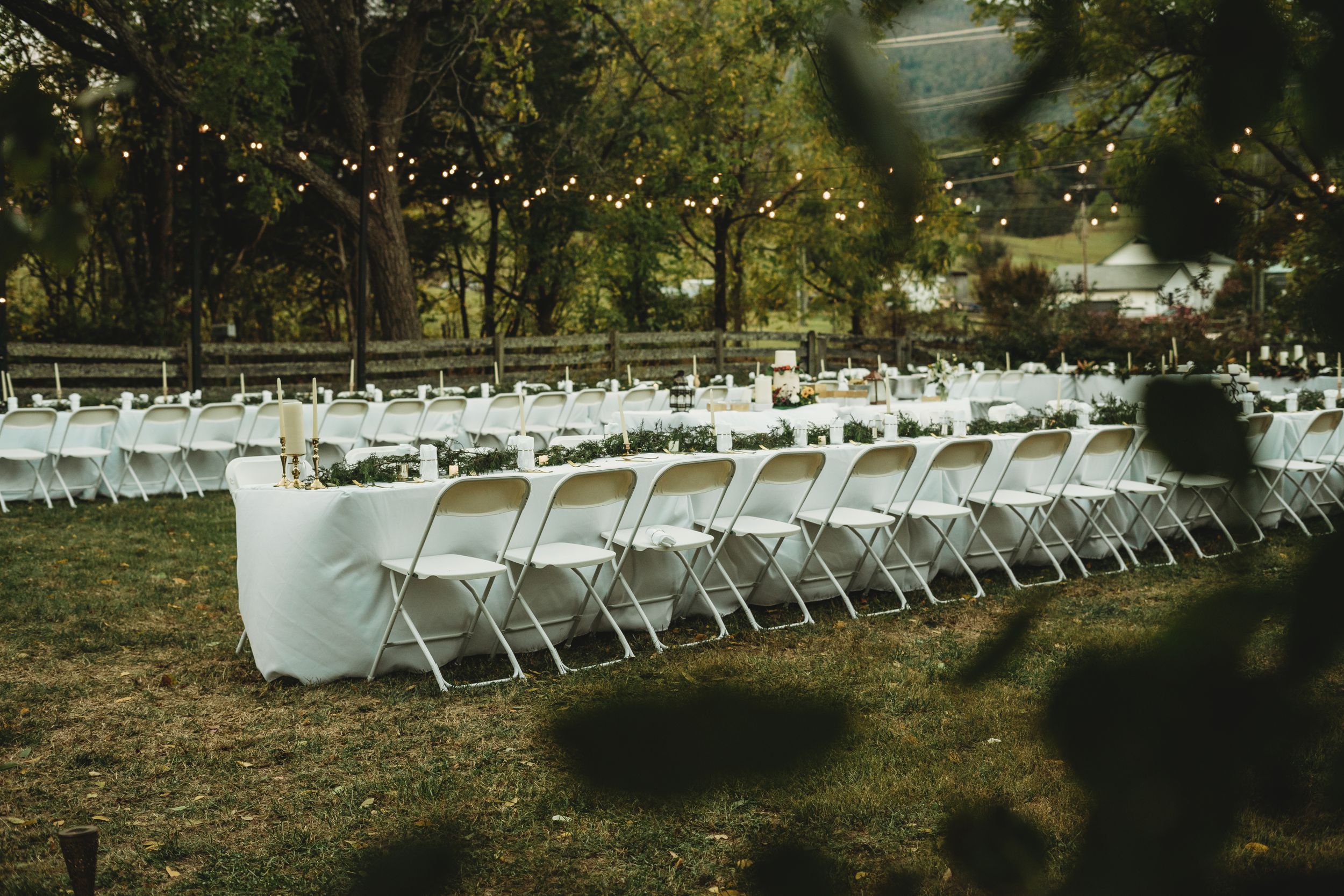 Rows of white folding chairs set up outdoors with string lights and trees in the background for an outdoor event.