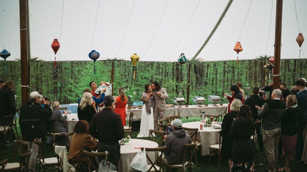 Panoramic view of wedding guests seated at tables under a tent in a forest setting.
