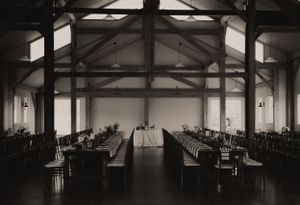 Black and white photo of an empty reception hall with exposed beam ceiling and formal table settings.