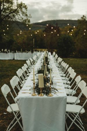 Long banquet table with white linens and candle centerpieces set up outdoors under string lights at dusk.
