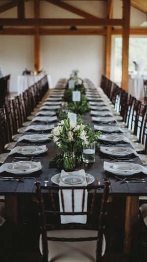 Elegant table setting with white plates and greenery centerpieces in a rustic wooden beam venue.