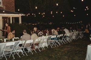 Evening outdoor dining setup with string lights illuminating rows of white folding chairs and tables.