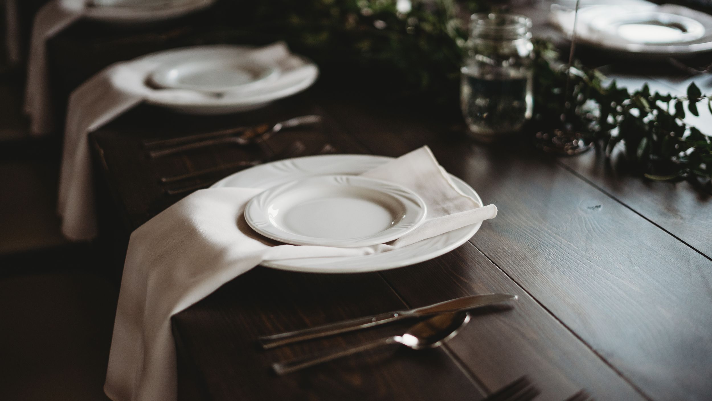White dinner plates and place settings arranged on rustic wooden tables for a wedding reception.