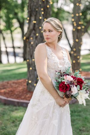Elegant bridal portrait with lace gown and burgundy bouquet against twinkling lights.
