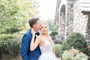 A romantic wedding portrait of a couple embracing in front of a stone building with lush greenery in the background.