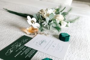 Wedding invitation with green details, perfume bottle, and white floral accents on textured surface.