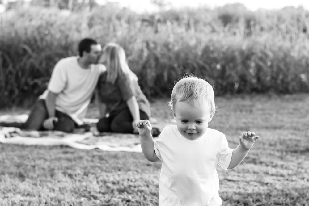 Black and white candid of joyful toddler playing while parents relax on blanket in background.