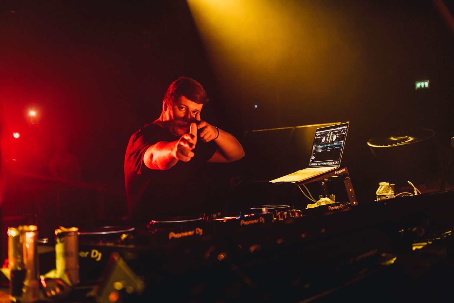 DJ performing at a nightclub under dramatic red and yellow stage lighting with DJ equipment and laptop.