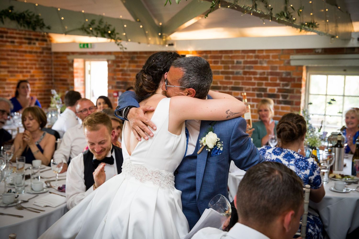 Wedding guests share joyful moments during an indoor reception with twinkling lights and exposed brick walls.