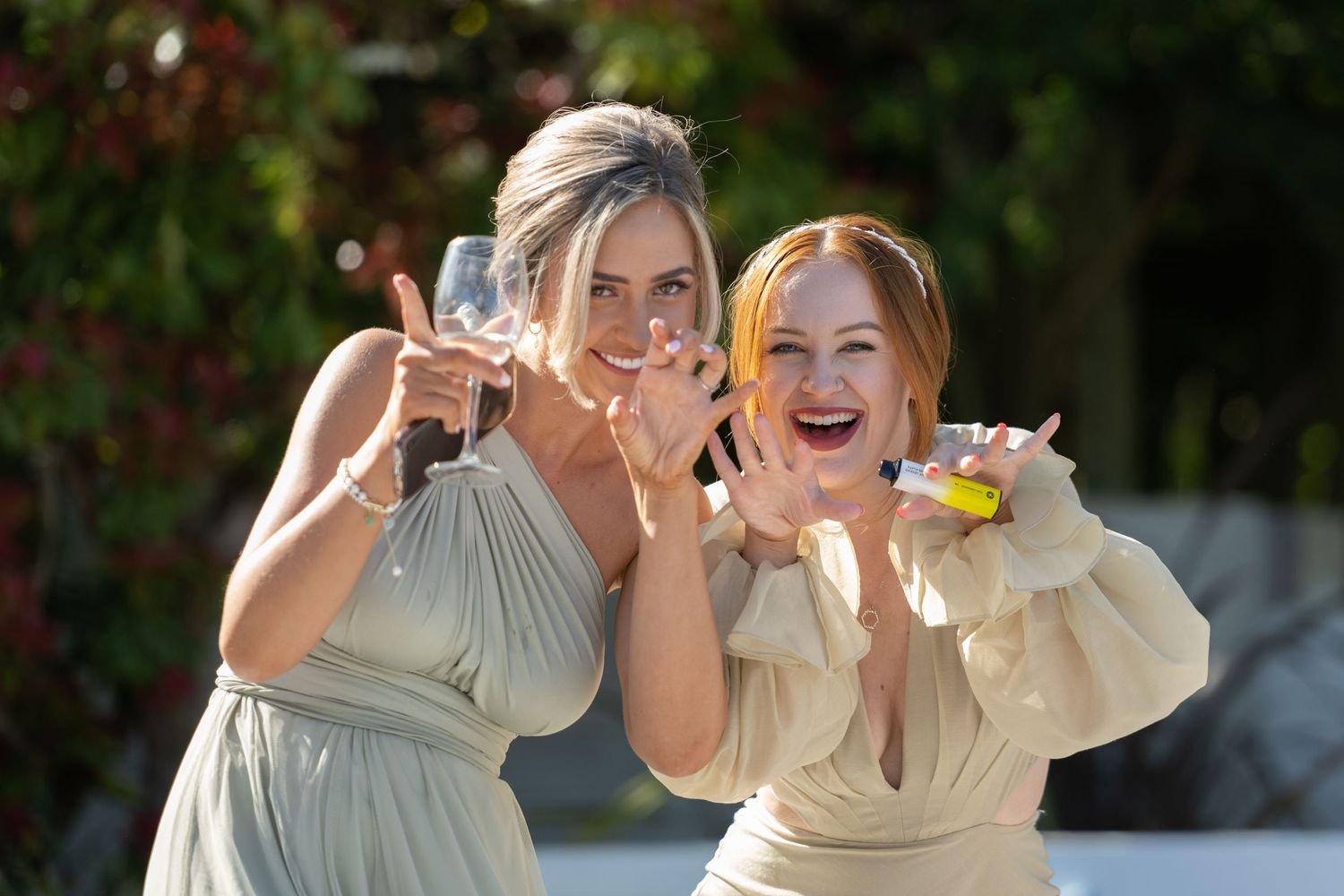 Two bridesmaids in light colored dresses laugh and pose together outdoors.