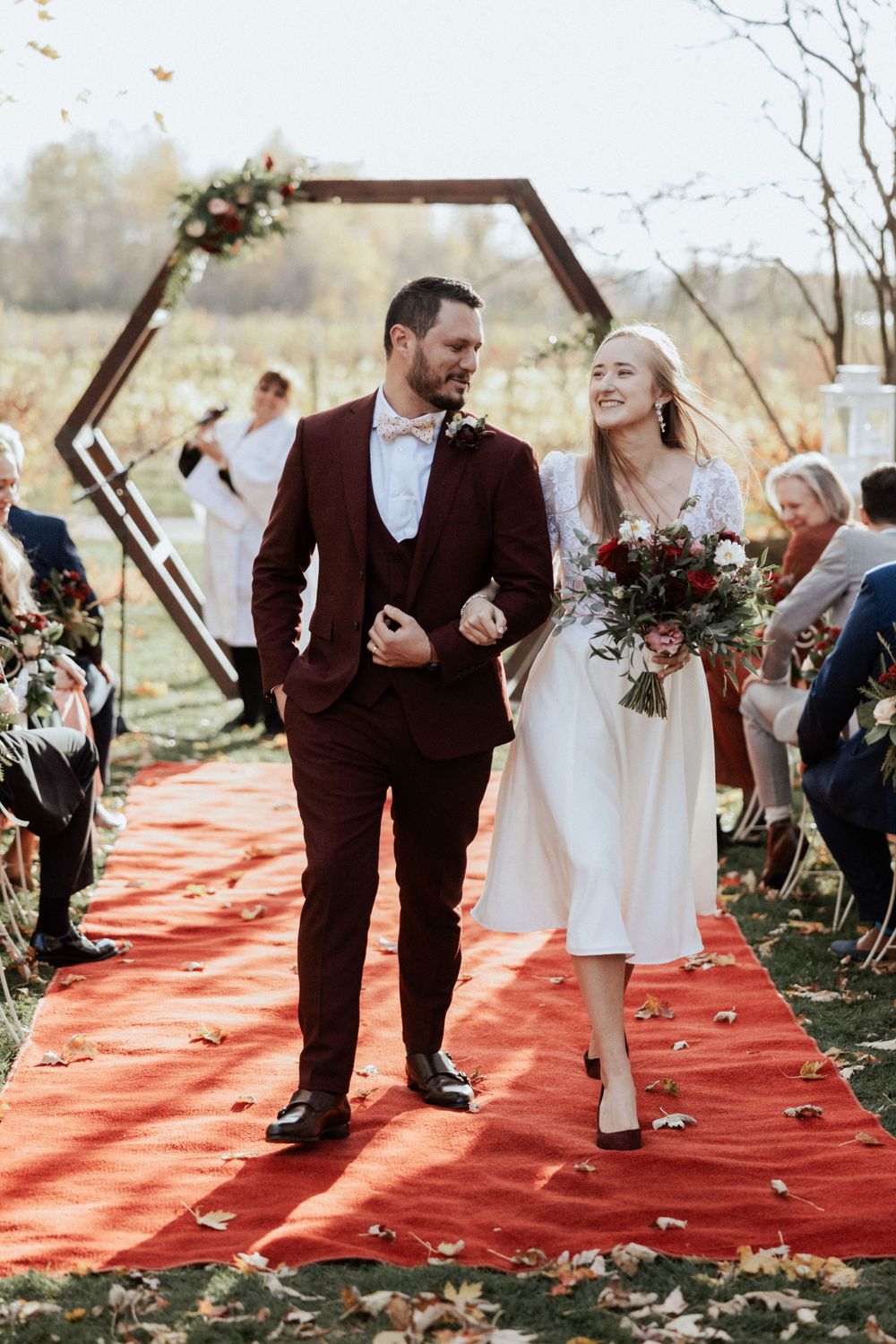 A couple walks down a red carpet aisle after their wedding ceremony under a geometric arch.