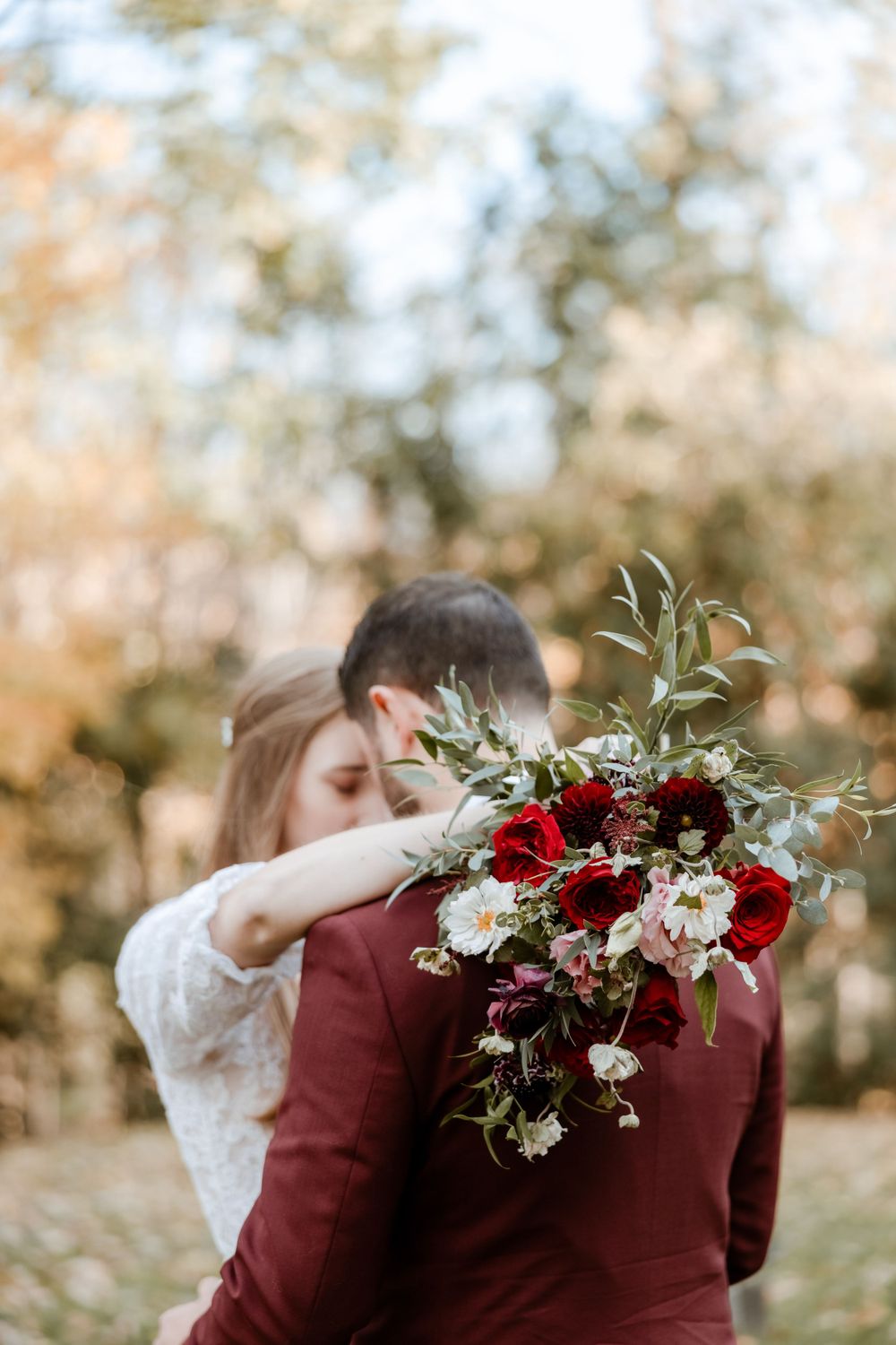 A romantic embrace captured from behind with a burgundy and white floral bouquet.
