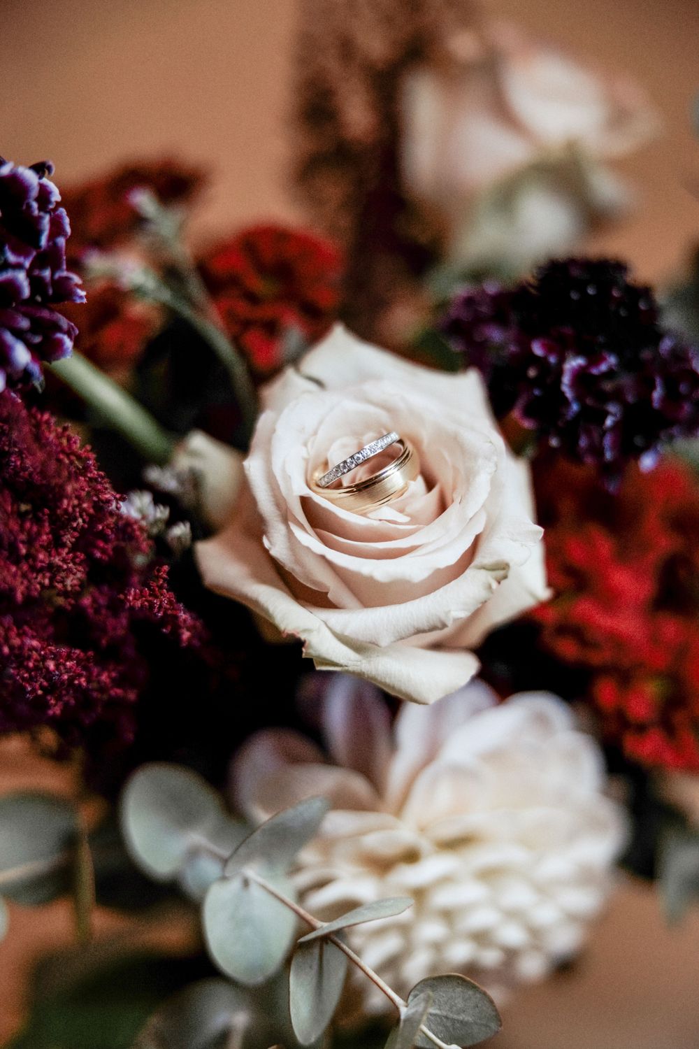 Wedding rings nestled in a blush pink rose surrounded by deep burgundy and purple flowers.