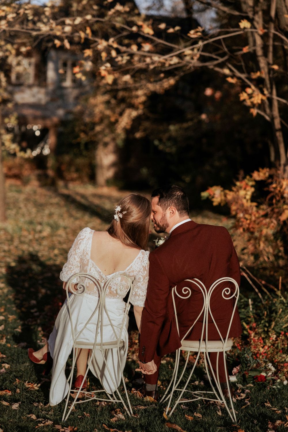 Newlyweds sitting on vintage white chairs surrounded by fall foliage at sunset.
