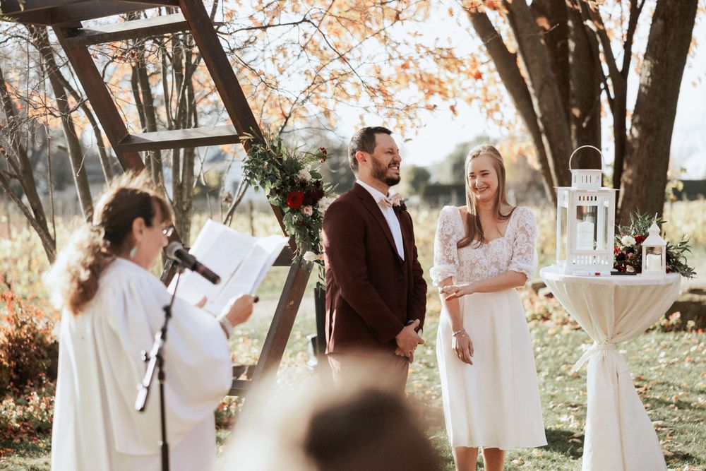 A wedding ceremony taking place outdoors with a wooden geometric arch and autumn foliage.