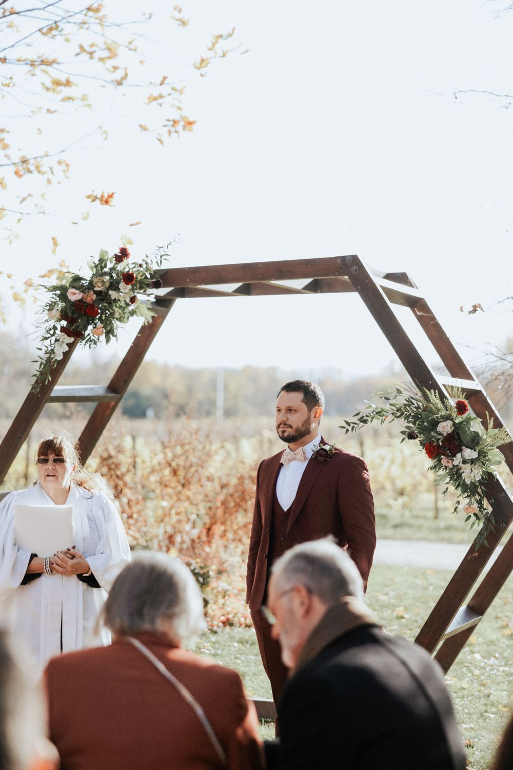 An outdoor wedding ceremony under a wooden hexagonal arch decorated with fall florals.