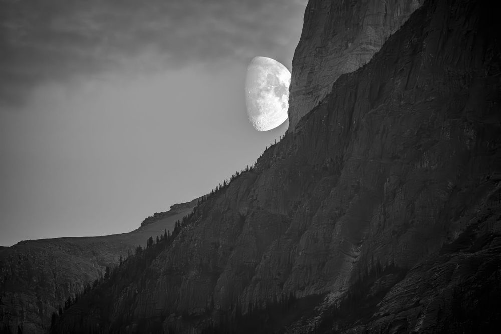 Large moon rising beside dramatic mountain cliff face in black and white.
