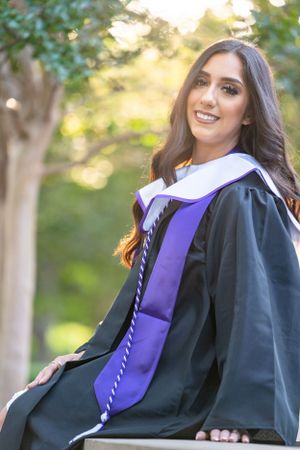 Graduate in academic regalia sits outdoors with natural sunlit background for graduation photo.