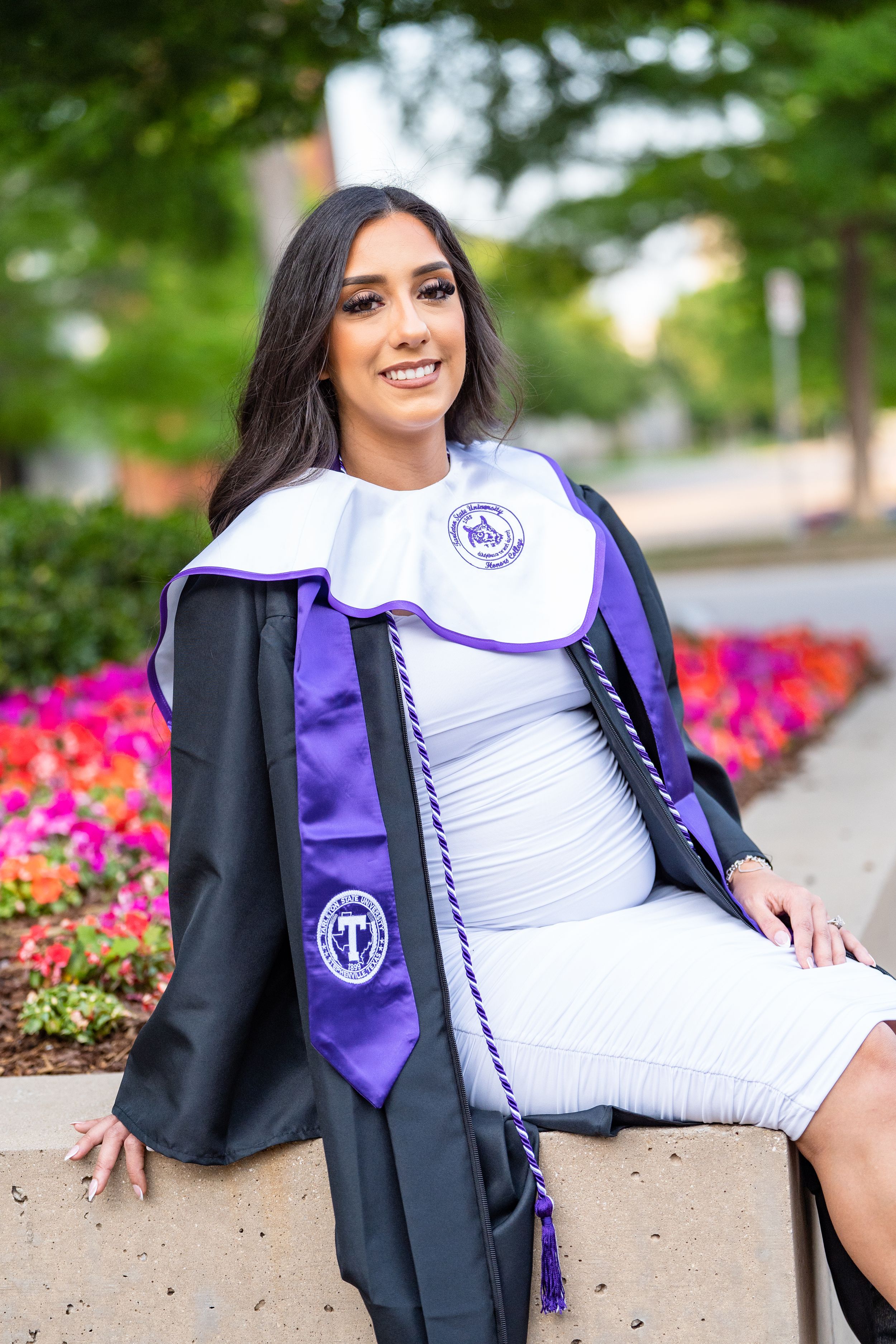 Graduate in black gown and purple stole poses by colorful flower bed for graduation portrait.