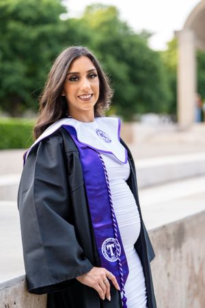 Graduate wearing black academic gown and purple stole poses outdoors in white dress for graduation photos.