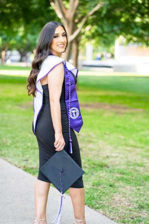 Graduate wearing black dress and purple academic regalia poses outdoors on university grounds.