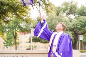Graduate wearing purple academic regalia poses outdoors on a sunny day at Texas Christian University.