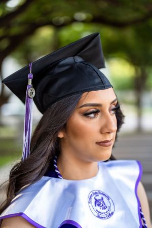 Side profile view of graduate wearing black cap with purple tassel against blurred green background.