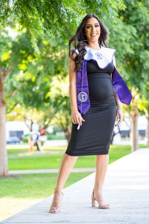 Graduate in black dress and purple graduation stole poses on campus walkway.