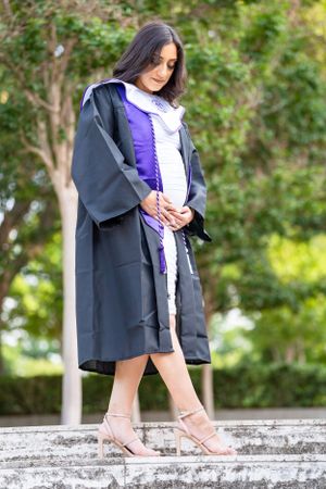 Graduate in full academic regalia looks down while standing on stone steps outdoors.