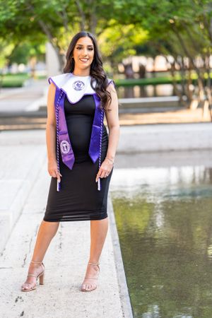 Graduate in black dress and purple stole poses near reflecting pool on campus.