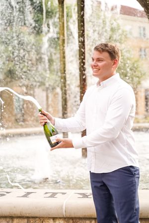 Graduate in formal attire pops champagne bottle in outdoor celebration.