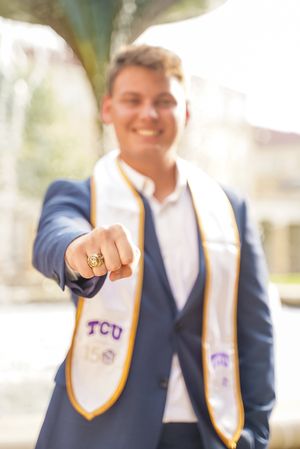 Graduate in TCU regalia shows off class ring and diploma outdoors.
