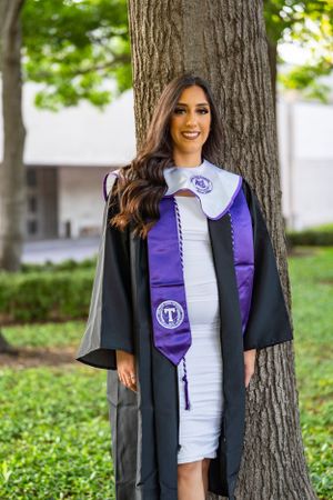 Graduate in black gown and purple stole poses by tree trunk for graduation portrait.