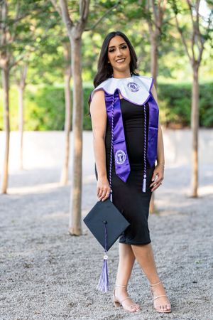 Graduate in black dress and purple regalia poses with mortarboard on tree-lined path.