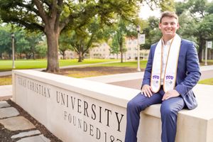 Graduate in blue suit sits on stone wall with university name engraved behind.