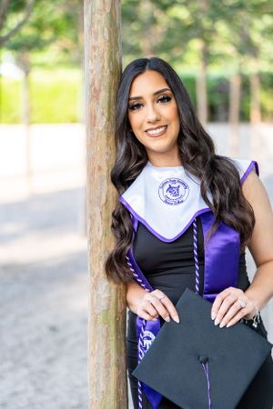 Graduate in academic regalia poses next to tree trunk in outdoor setting.