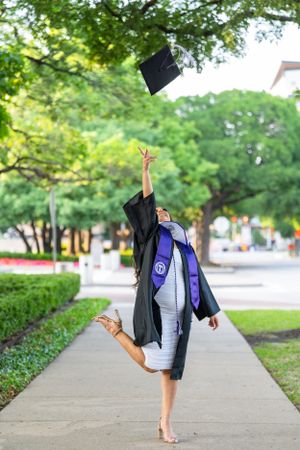Graduate throws cap in celebratory pose while balancing on one foot outdoors.